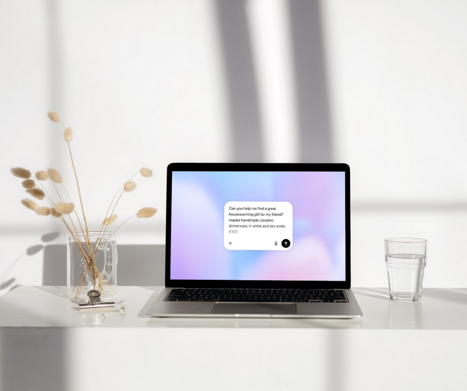 A laptop sits on a white desk in soft natural light, displaying a ChatGPT-style conversation bubble that reads: “Can you help me find a great housewarming gift for my friend? Maybe handmade, ceramic dinnerware, in white and tan under $100.” A glass of water and a minimalist vase with dried flowers are beside the laptop, creating a clean, modern workspace aesthetic.